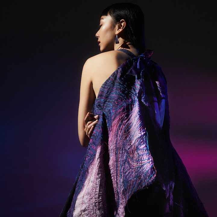 Frau mit schimmerndem, lila Kleid, blickt über die Schulter in ein düsteres Licht.