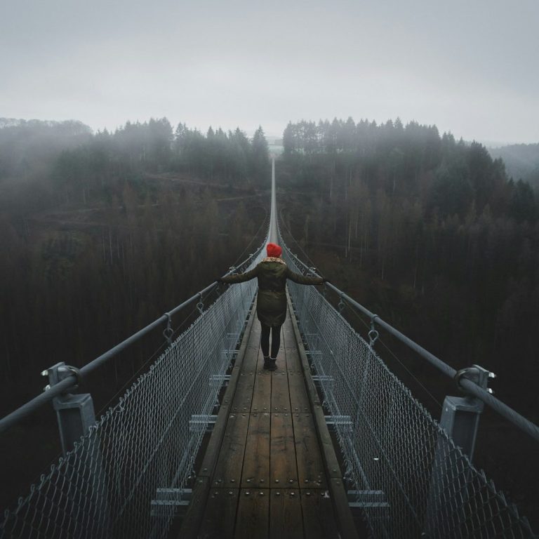 Person mit rotem Hut steht auf einer Hängebrücke in nebliger Landschaft.